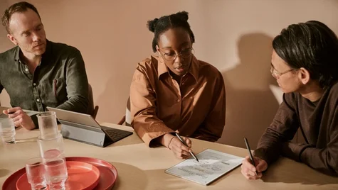 Three people sitting by a table, engaging in a discussion while pointing at a reMarkable Paper Pro lying on the table.